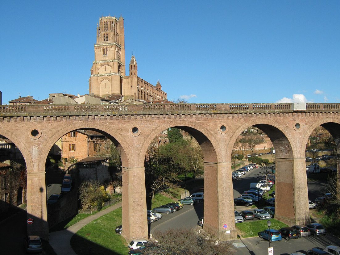 Albi Cathedral, a vast red-brick Gothic church with a tall square bell tower, seen above the arches of a brick viaduct