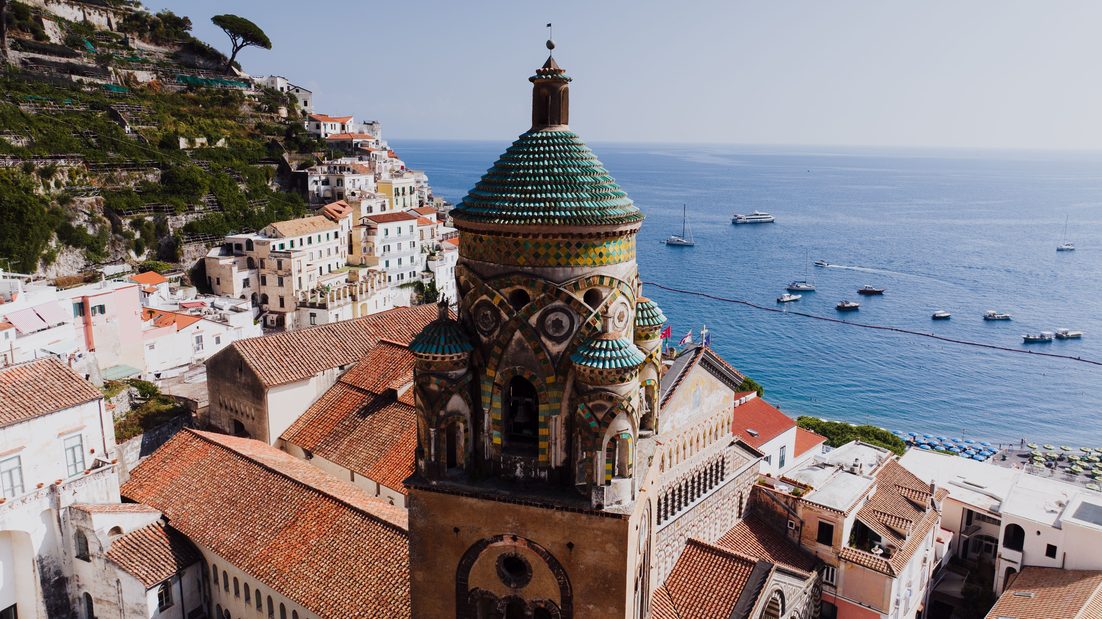The Romanesque bell tower of Amalfi Cathedral with its green and yellow tiled crown, above the rooftops of Amalfi and the blue sea