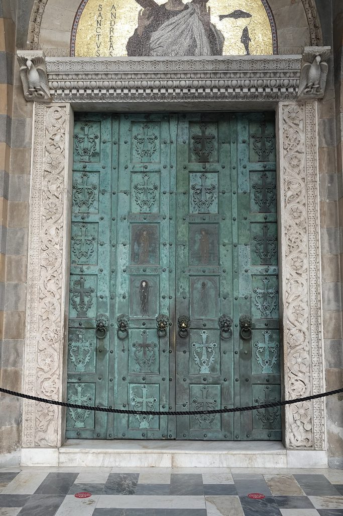 The green bronze doors of Amalfi Cathedral, divided into panels, under a carved marble frame and a mosaic of St Andrew