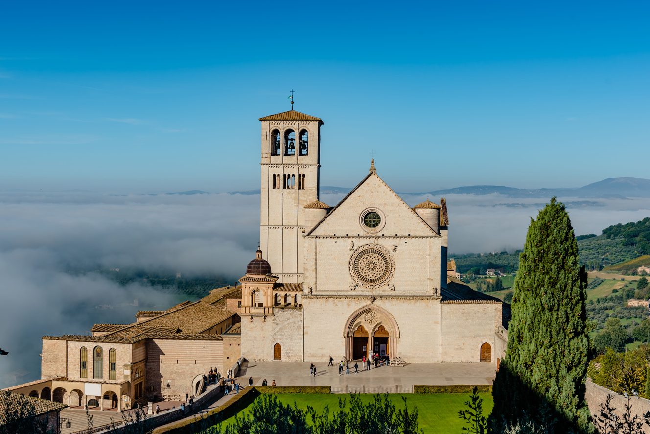 The plain white stone facade and rose window of the Upper Basilica of St Francis, with mist in the valley beyond