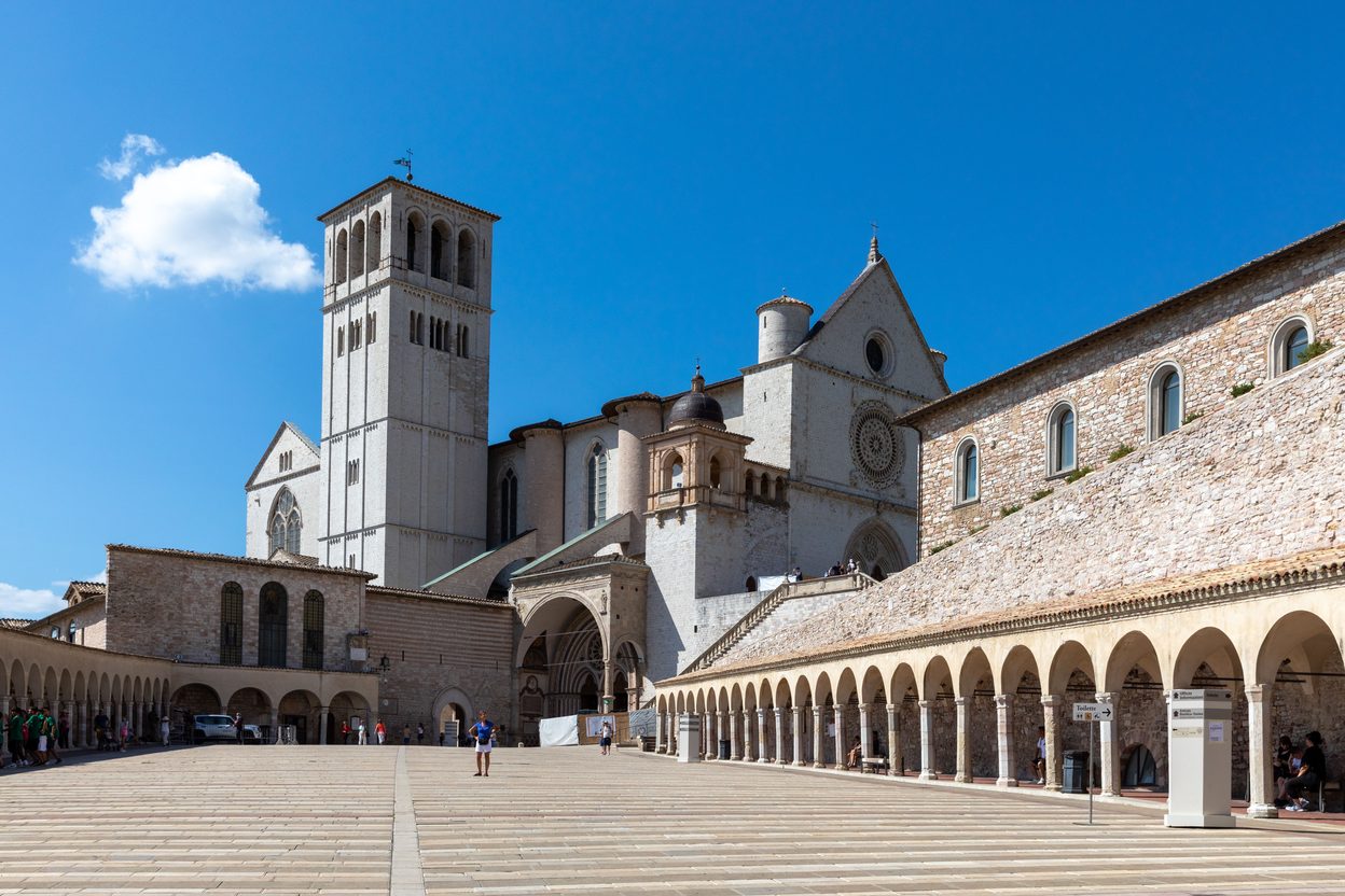 The Basilica of St Francis in Assisi, with its bell tower and the arcades of the lower piazza under a blue sky