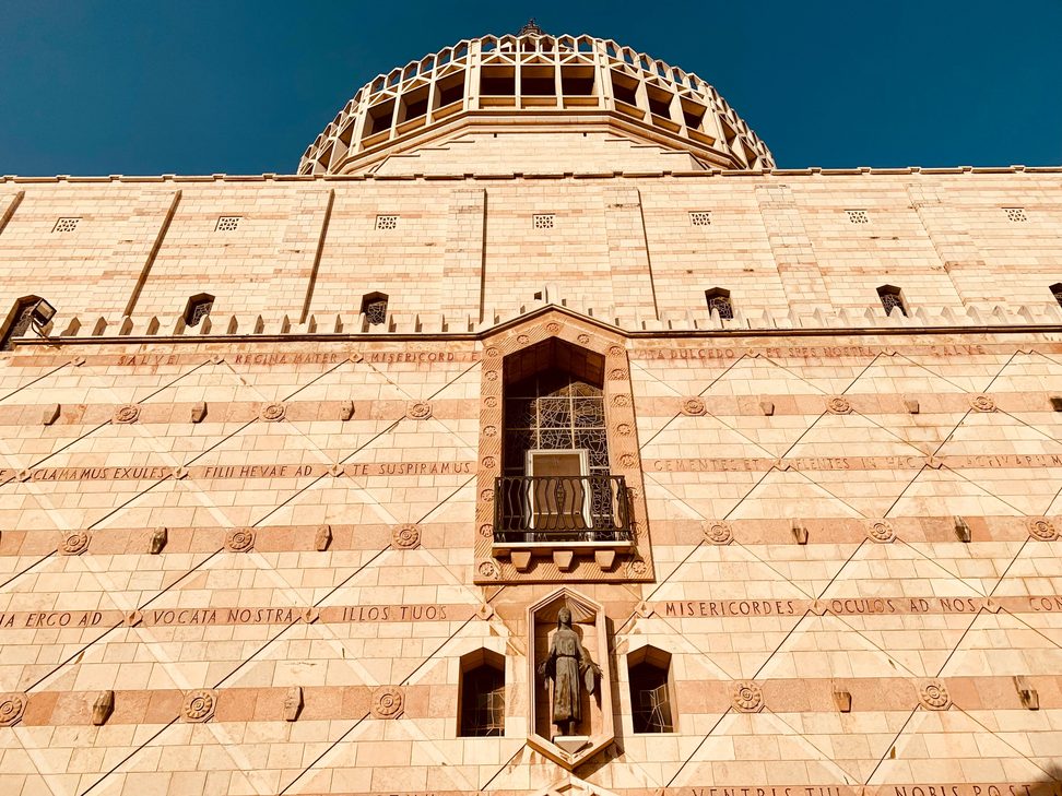 The pale stone west front of the Basilica of the Annunciation in Nazareth, with its tall window, reliefs and the dome behind