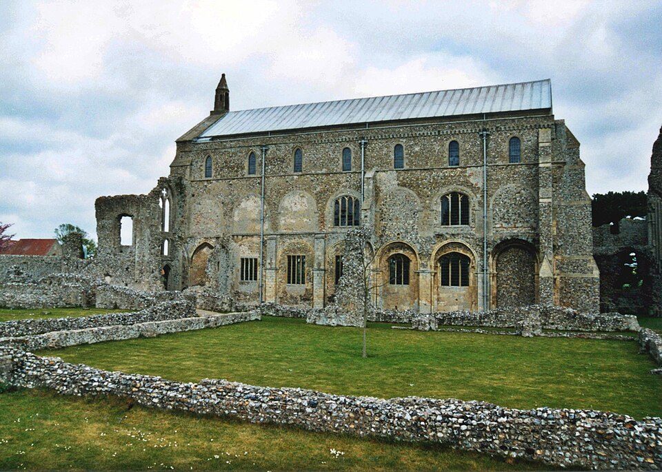 The long flint and stone south side of Binham Priory church with the ruined walls of the cloister in the grass in front