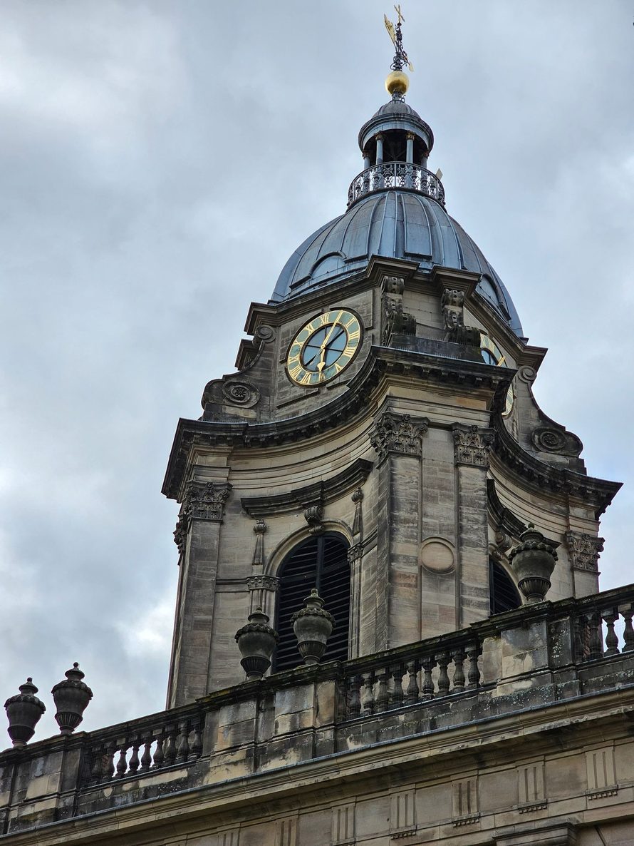 The baroque tower of Birmingham Cathedral, with its clock, curved sides and small dome, against a cloudy sky