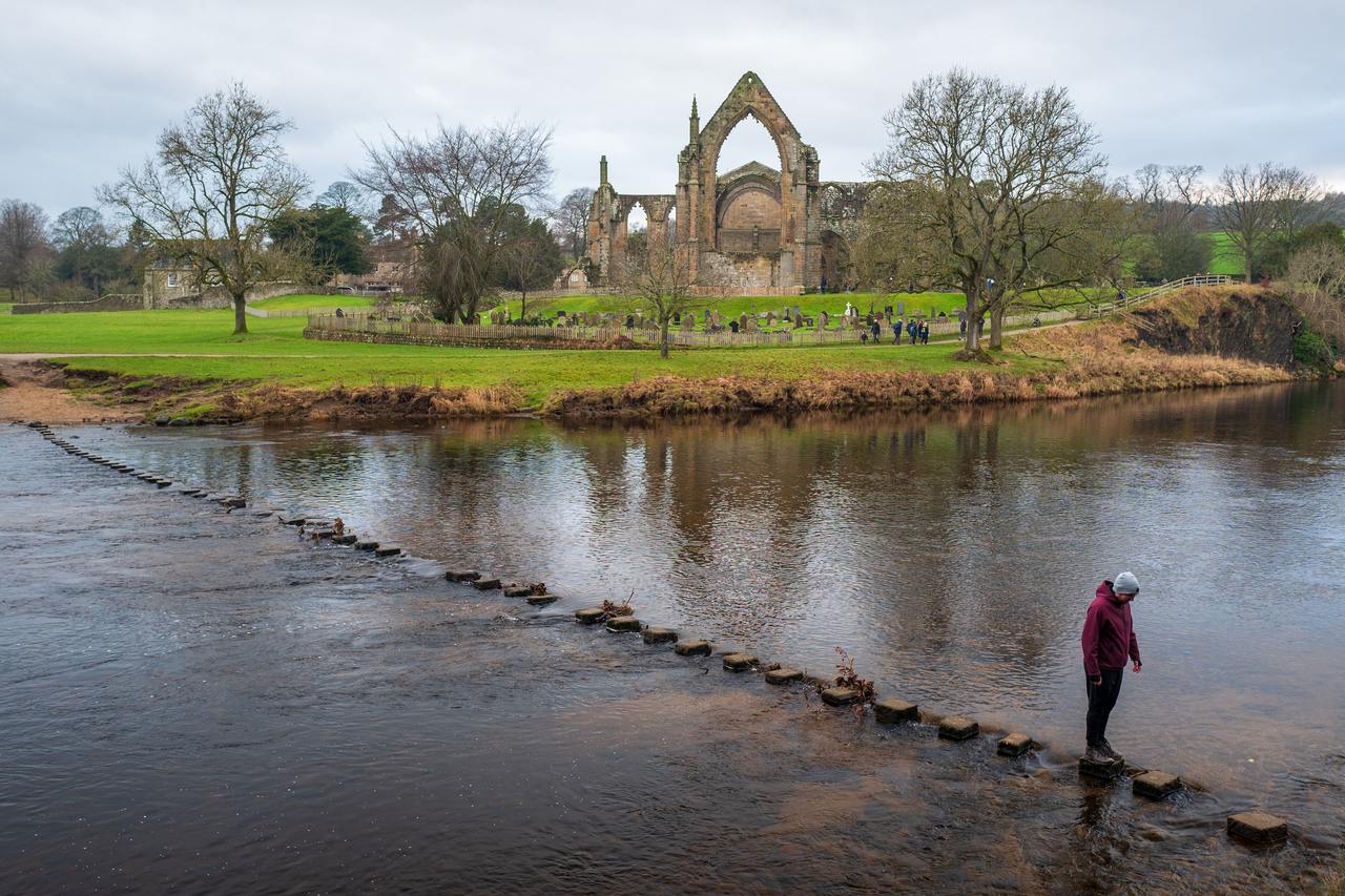 A walker crossing the stepping stones over the River Wharfe below the ruins of Bolton Priory