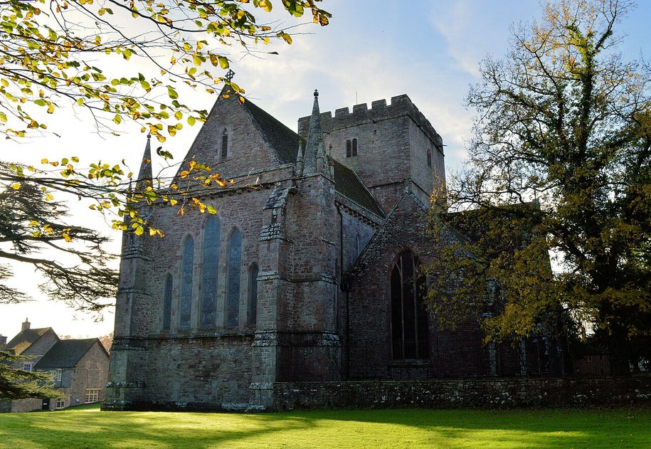 Brecon Cathedral seen across the lawn, its long choir and transept gable with the square central tower behind, framed by trees