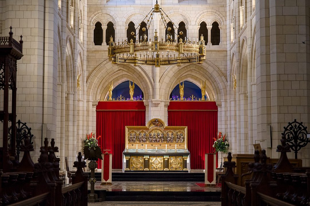 The gilded high altar of Buckfast Abbey under a large crown-shaped chandelier, with red hangings behind