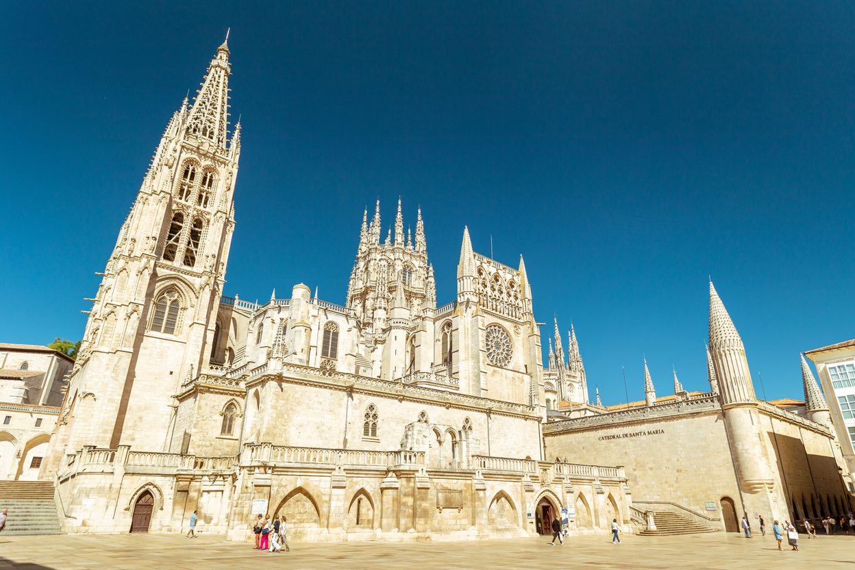The pale Gothic bulk of Burgos Cathedral with its openwork spires and pinnacles under a deep blue sky, seen from the square