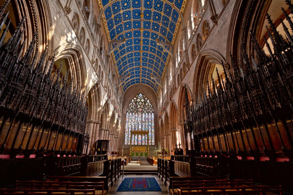 The choir of Carlisle Cathedral with dark carved stalls under a blue ceiling painted with gold stars, and the great east window