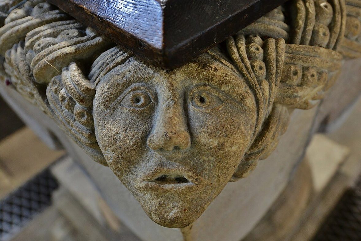 A carved stone head with staring eyes at the corner of the Norman font at Castle Rising