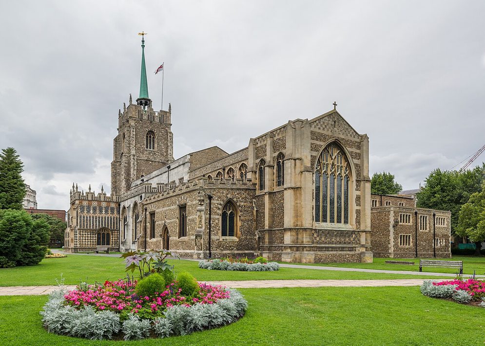 Chelmsford Cathedral seen across a lawn and flower bed, a long flint and stone church with a west tower and slender spire