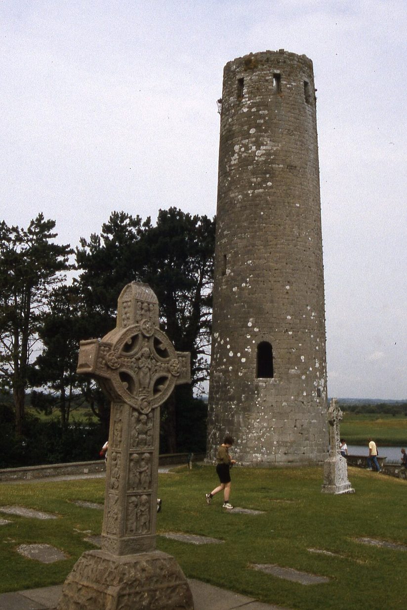 A ringed high cross carved with figure panels standing among grave slabs in front of O'Rourke's round tower at Clonmacnoise