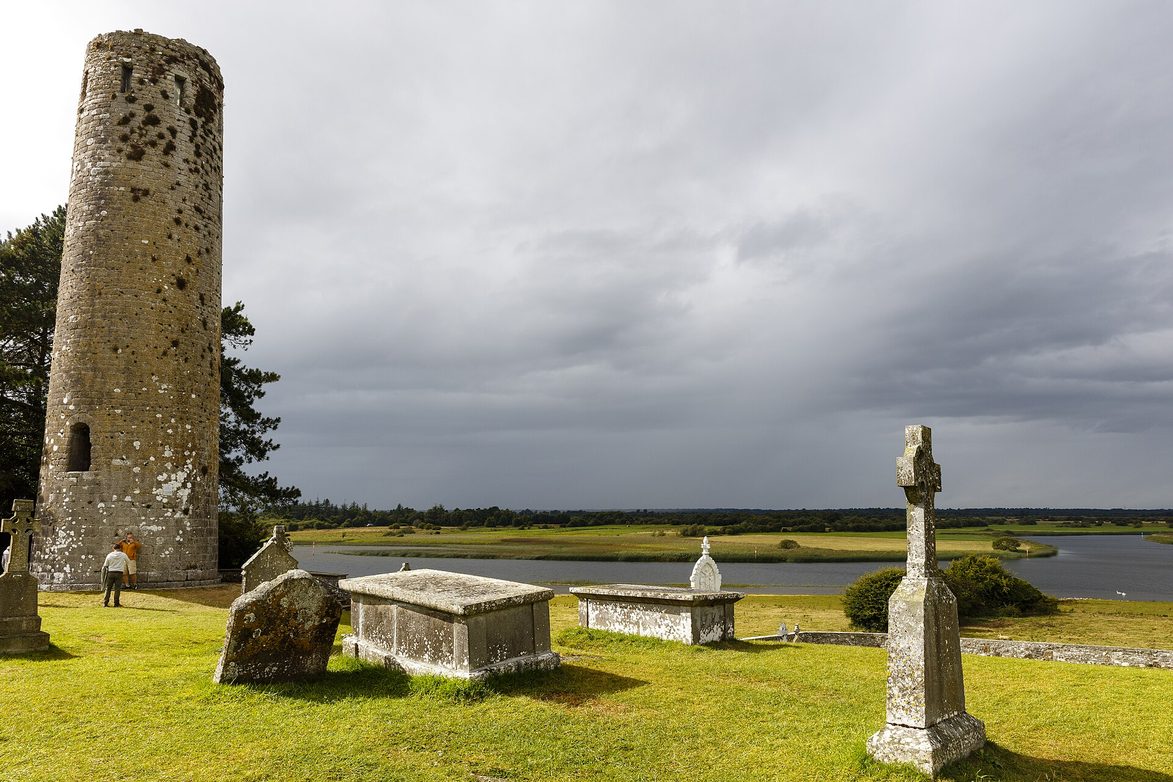 O'Rourke's round tower at Clonmacnoise beside a high cross and old graves, with the flat callows of the River Shannon behind