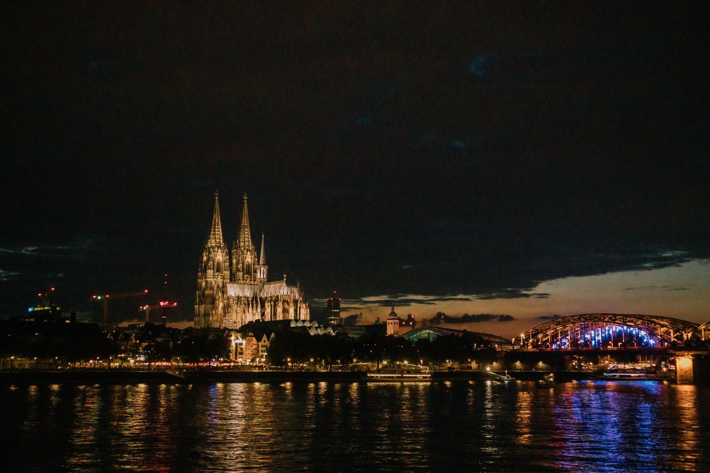 Cologne Cathedral lit up at night above the Rhine, with the lit Hohenzollern Bridge on the right