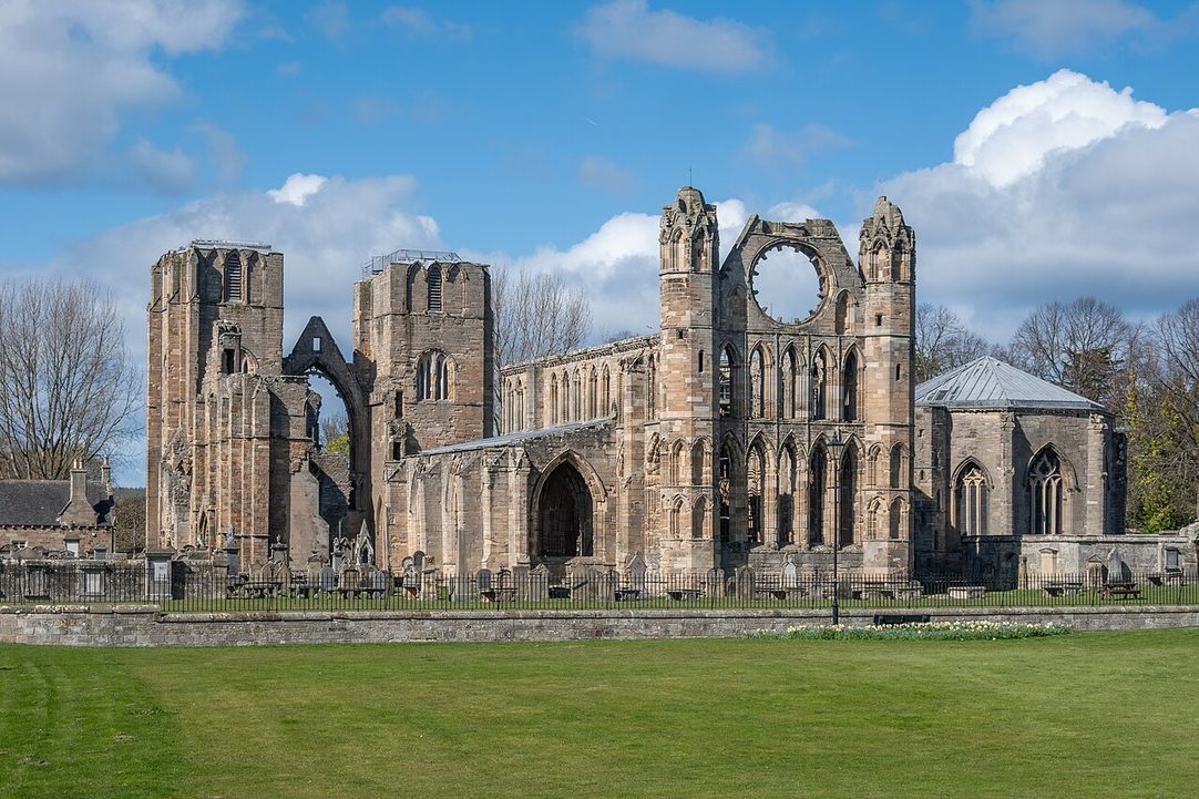 Elgin Cathedral ruins seen across a road and green from the north-east, with trees around