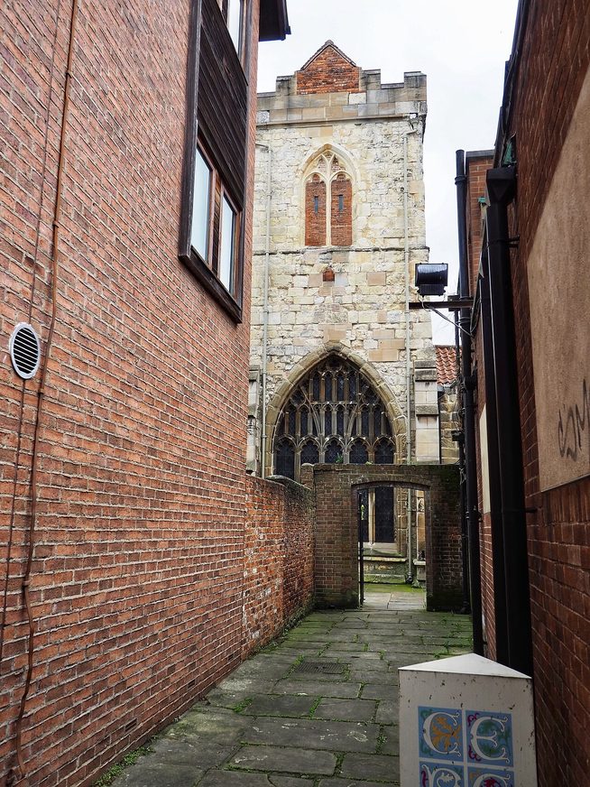 A narrow paved passage between brick walls leading from Goodramgate to Holy Trinity Church, York, with its tower ahead