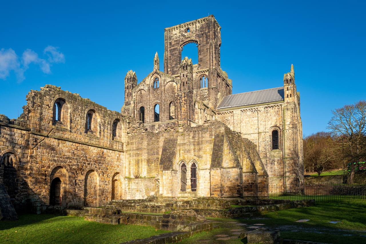 The sunlit ruins of Kirkstall Abbey with its broken crossing tower and the long walls of the church