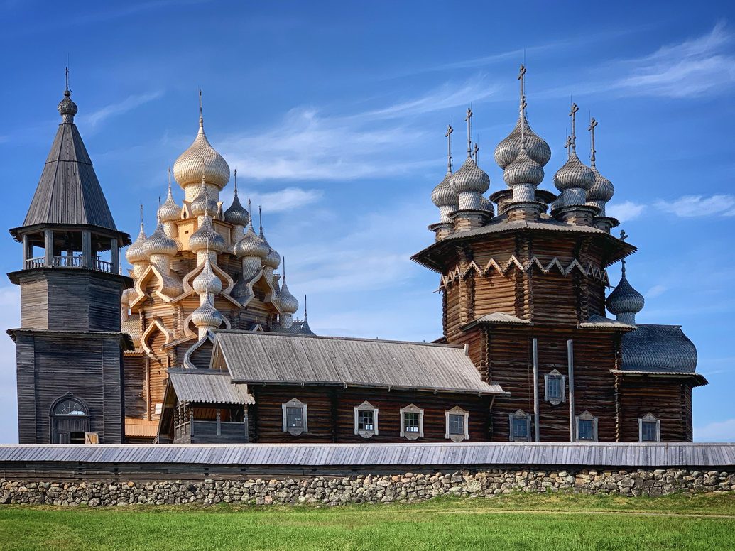 The three wooden buildings of the Kizhi Pogost inside their log fence on a grassy island under a blue sky