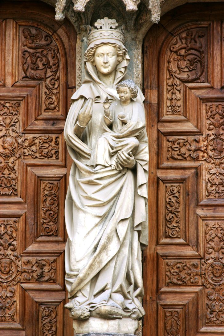 A white stone statue of the Virgin and Child on the central pillar of the west portal of León Cathedral, between carved wooden doors