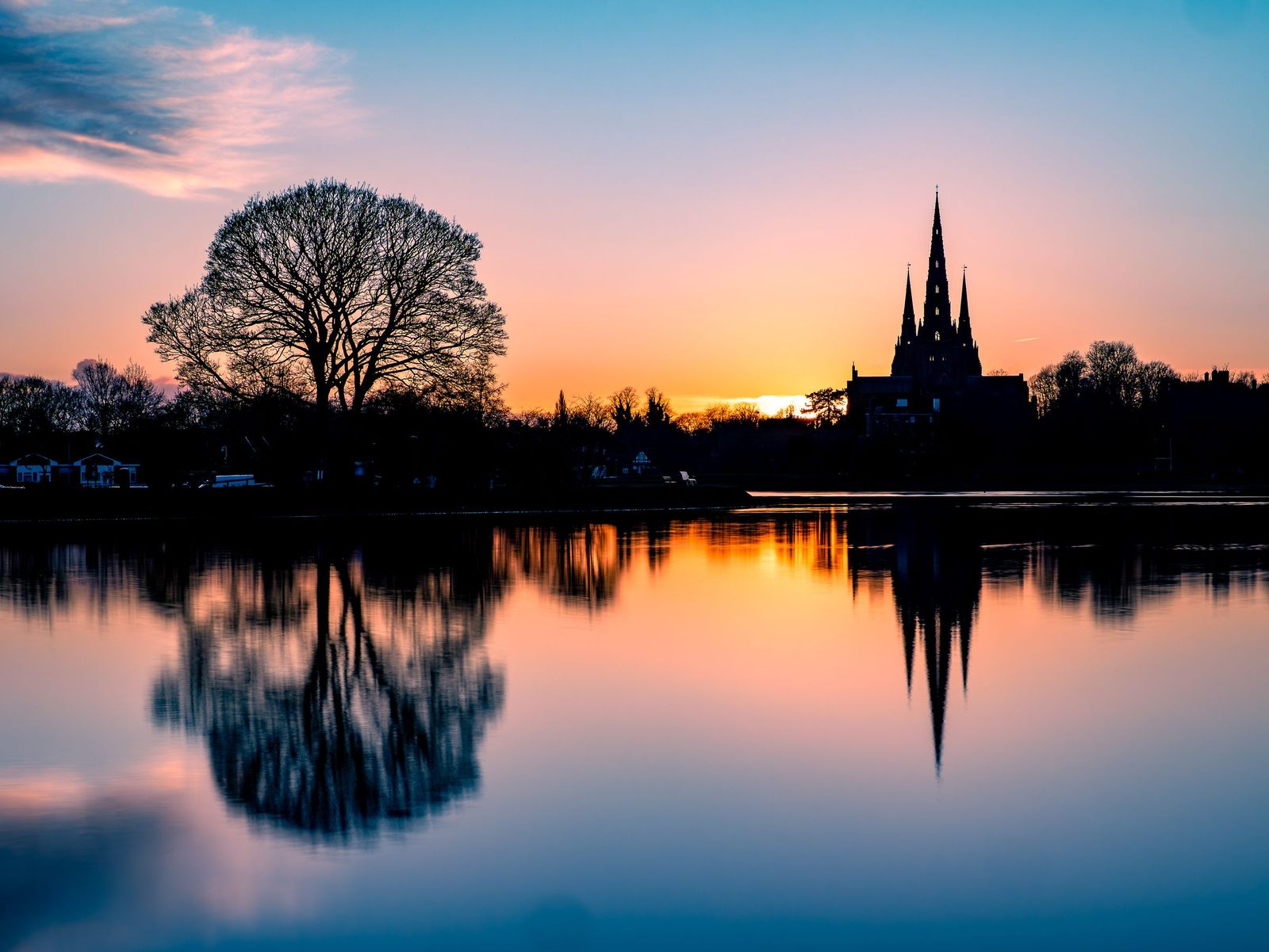 The three spires of Lichfield Cathedral silhouetted at sunset and reflected in Stowe Pool