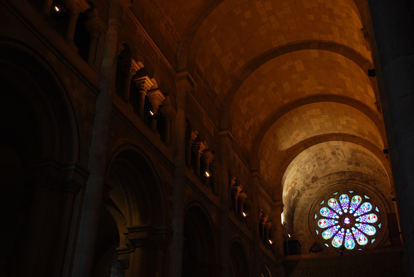 The dark Romanesque nave of Lisbon Cathedral, with round arches and the rose window glowing at the west end