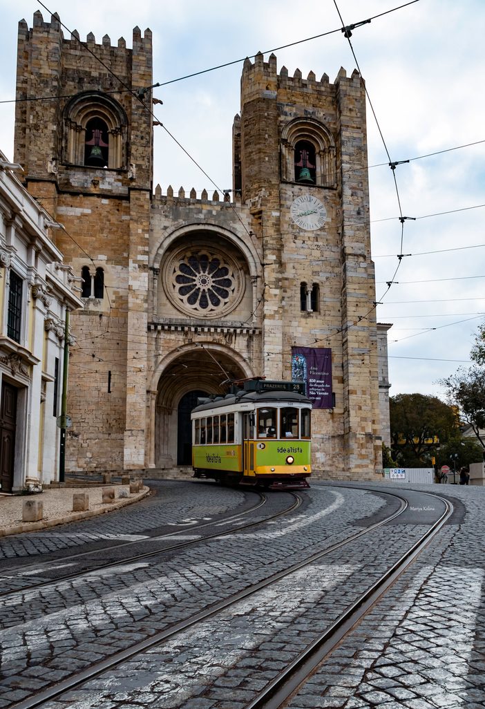 A yellow tram passing in front of the twin towers of Lisbon Cathedral on a cobbled street
