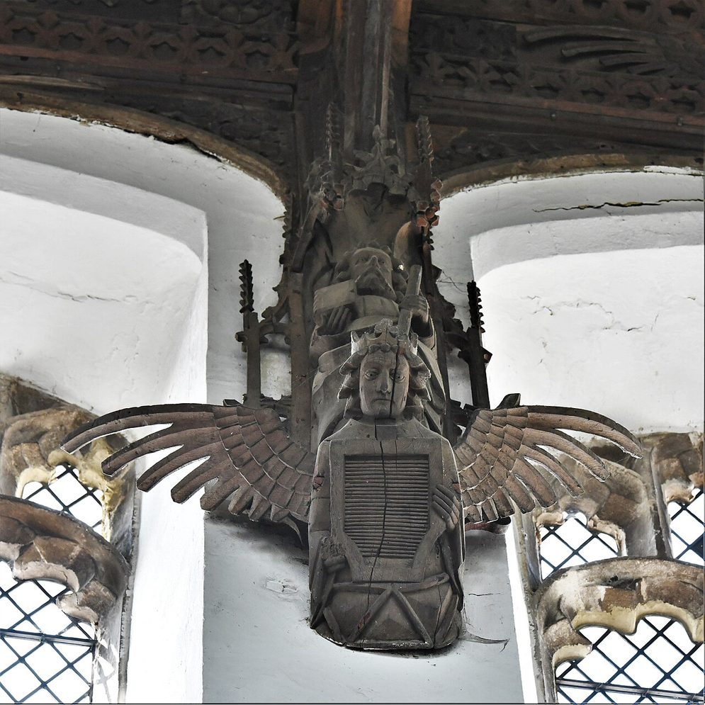 A carved wooden angel holding a shield below a saint in a niche, high on the wall of St Wendreda's, March