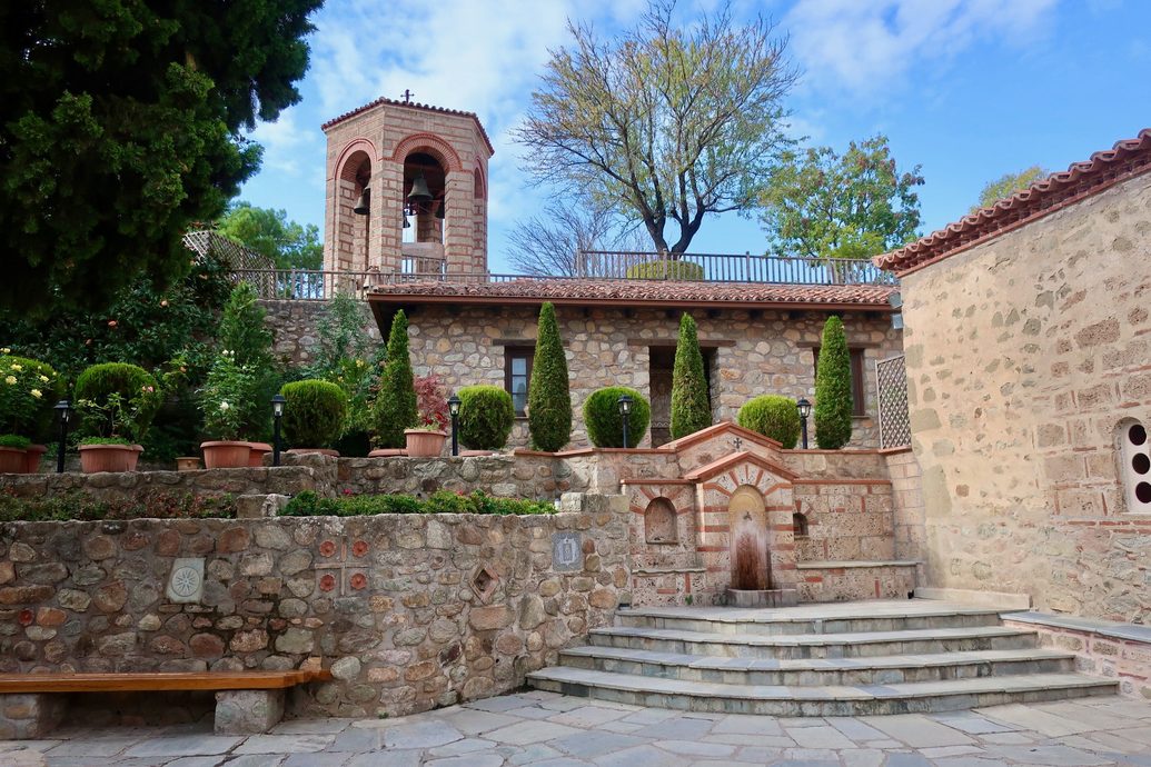 A stone courtyard at a Meteora monastery with steps, potted plants and a bell tower