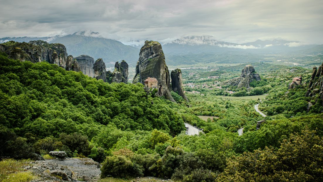 The grey rock pillars of Meteora rising above green woodland, with mountains on the horizon