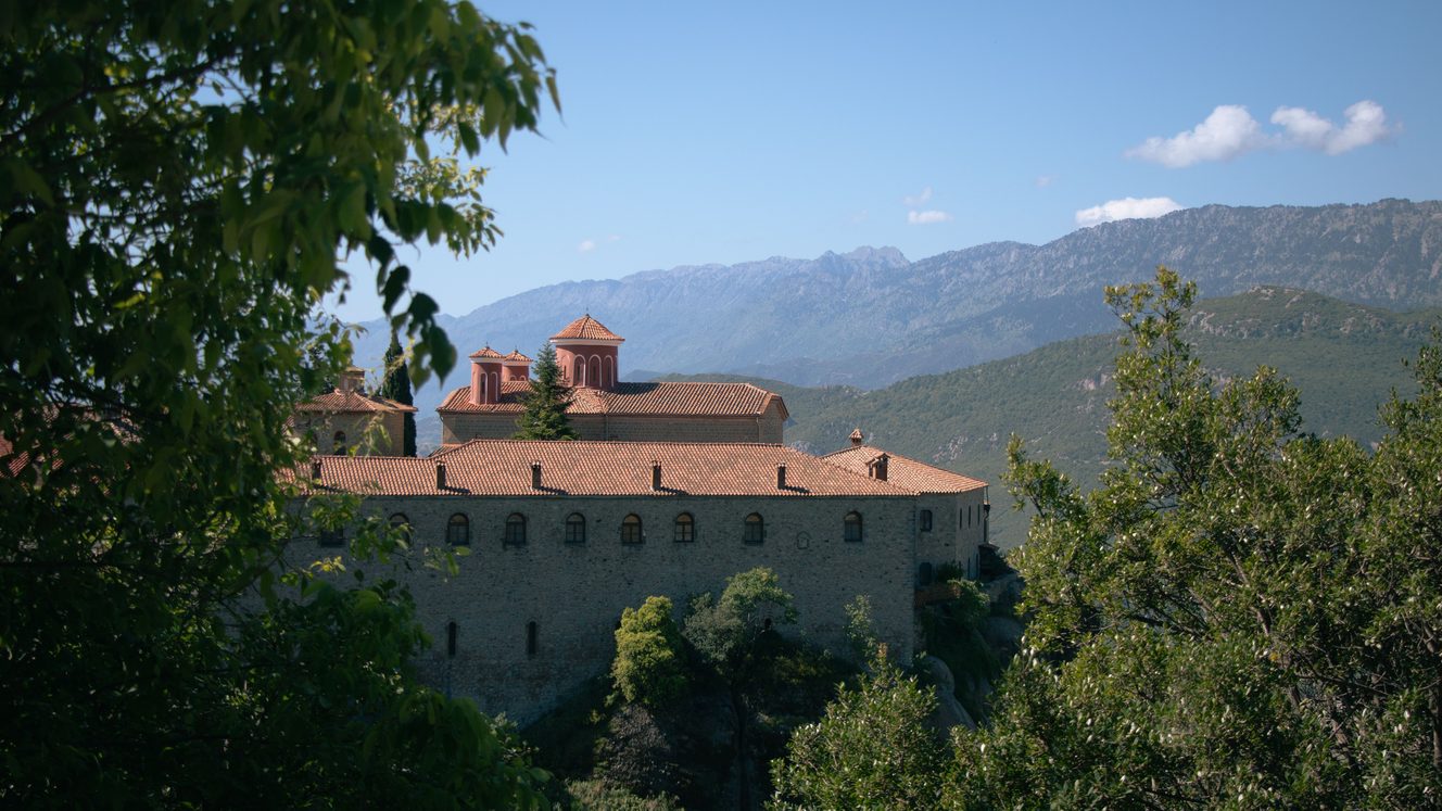 The long buildings of St Stephen's monastery at Meteora among trees, with mountains beyond