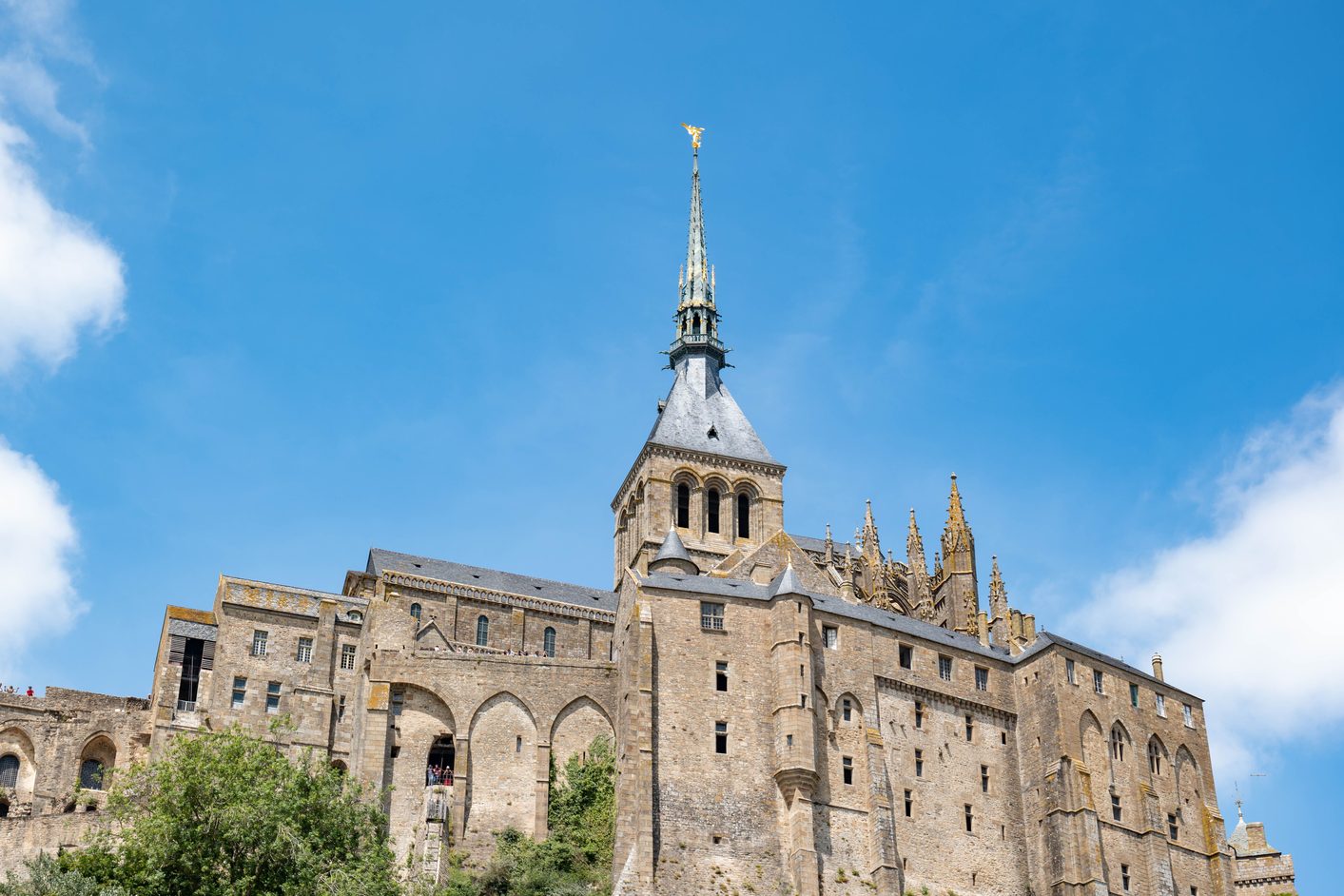 The Gothic abbey buildings and spire of Mont-Saint-Michel seen from below against a blue sky