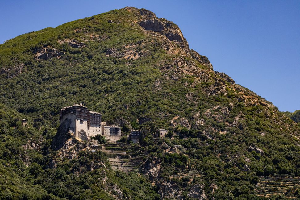 A stone monastery building on a steep wooded hillside of Mount Athos under a blue sky