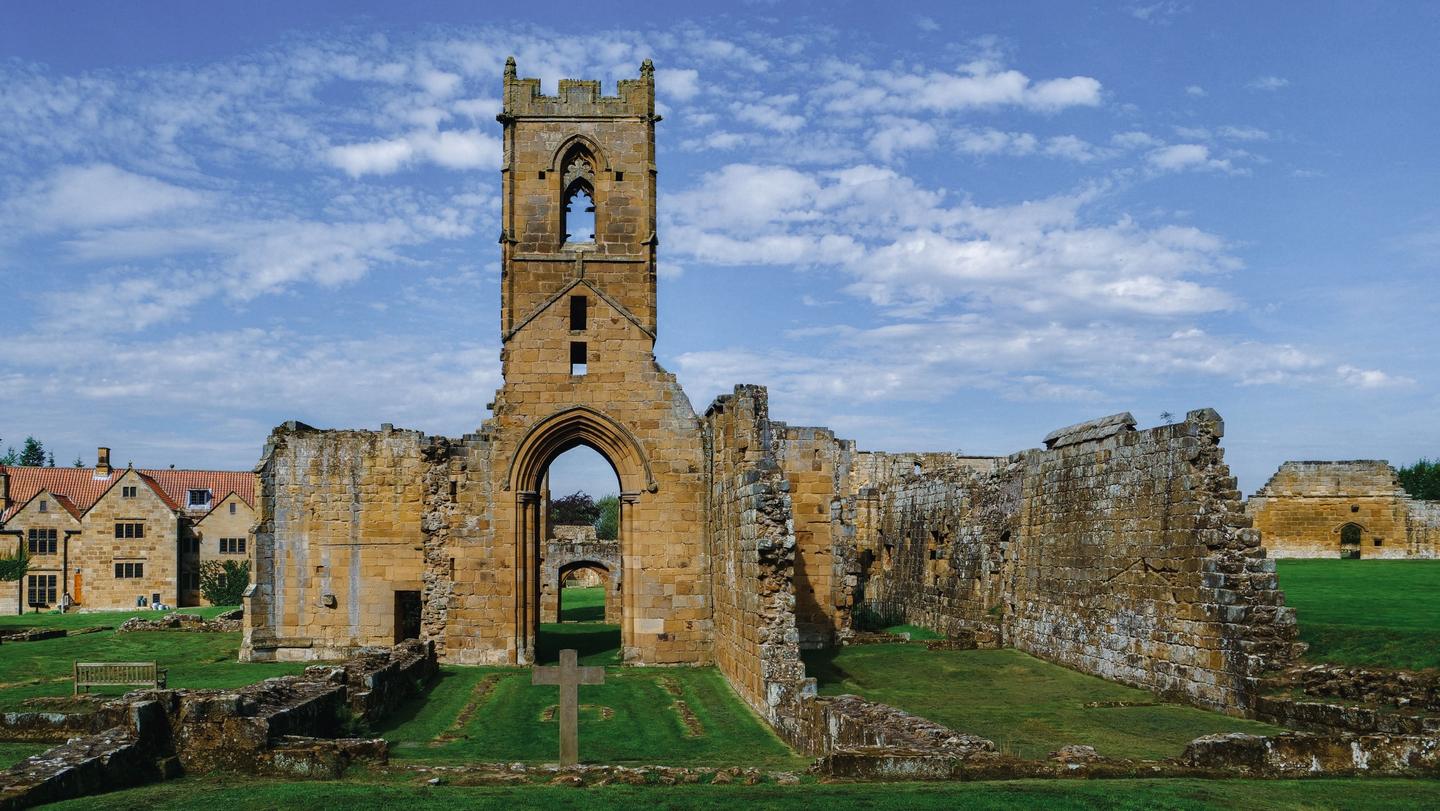 The ruined church and tower of Mount Grace Priory on a lawn under a blue sky, with the manor house to the left