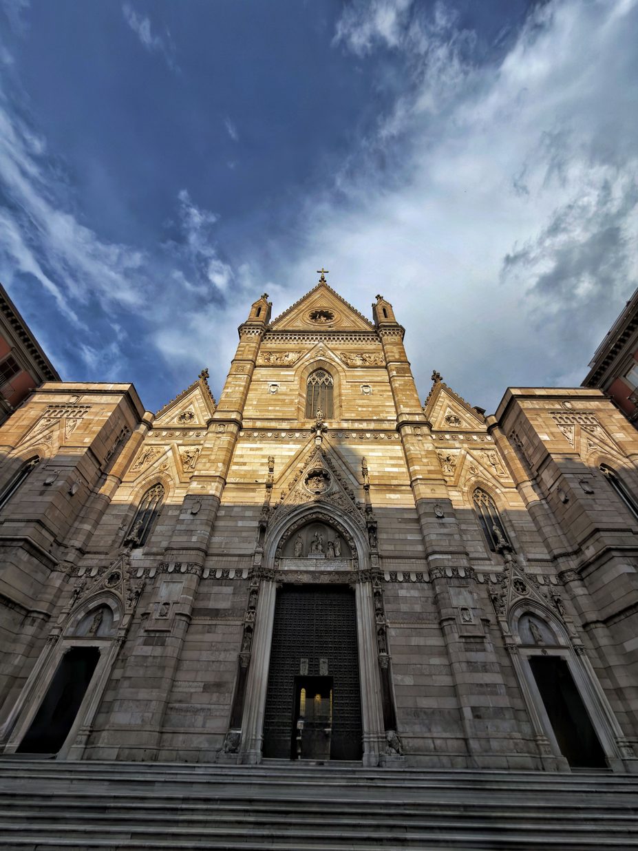 The white neo-Gothic facade of Naples Cathedral, with its three pointed portals and gabled centre, seen from below against a stormy sky