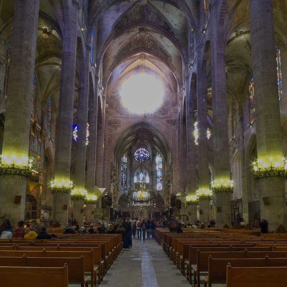 The immensely tall nave of Palma Cathedral, with slender columns, lit chandeliers and a glowing rose window at the east end