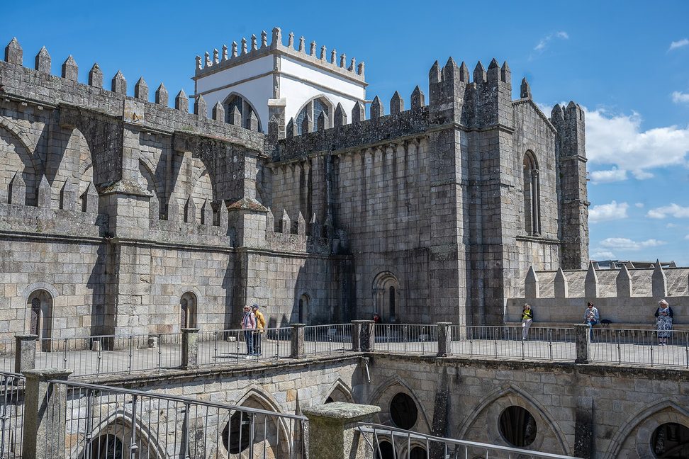 The battlemented granite walls and towers of Porto Cathedral rising above the terrace of its cloister, under a blue sky