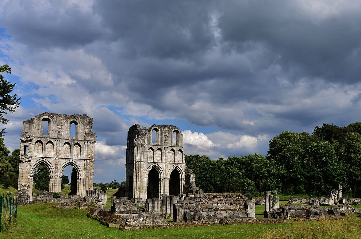 The low foundations of the monastic buildings at Roche Abbey on a lawn, with the transepts beyond under a stormy sky