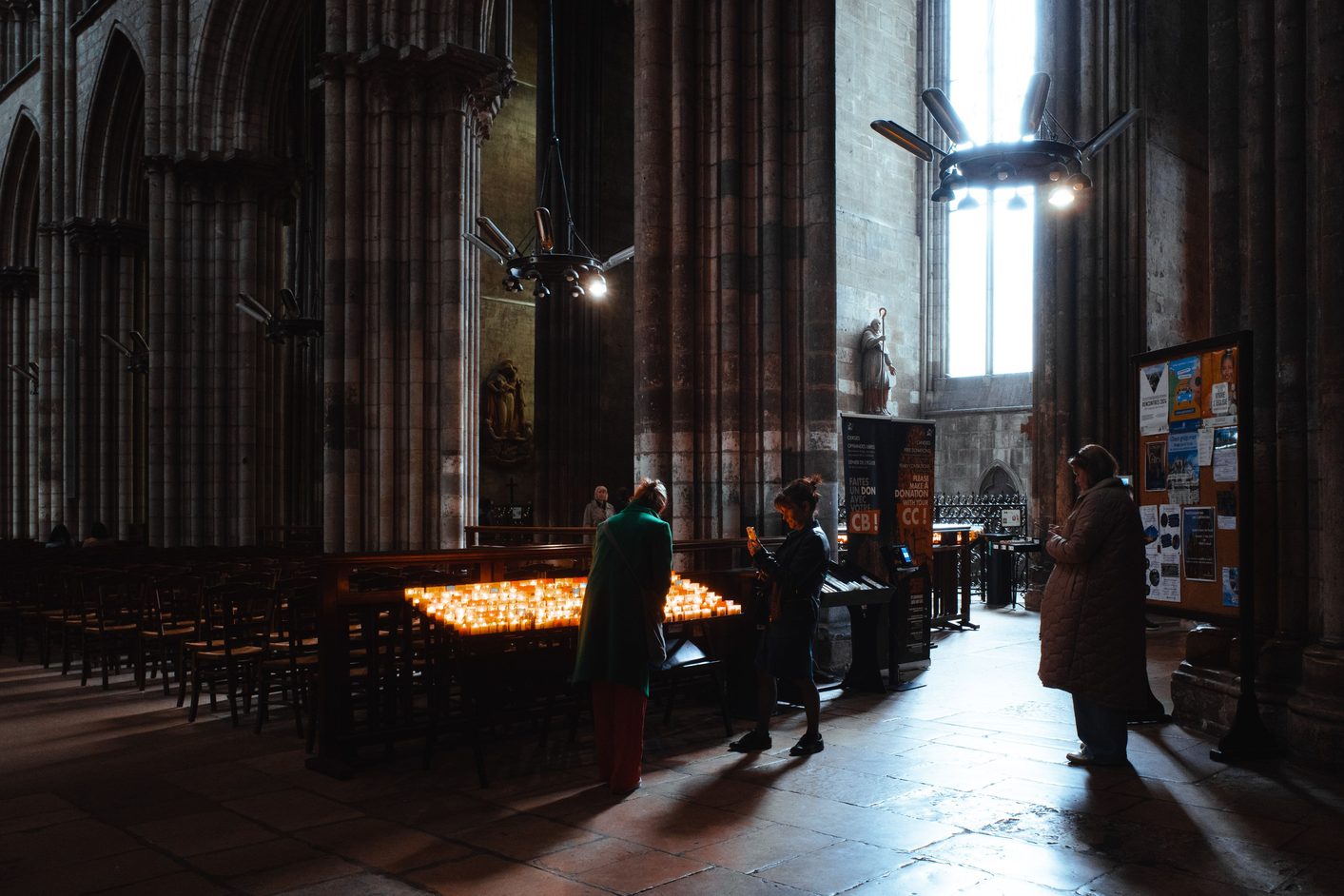 Visitors lighting candles beside a tall pier inside Rouen Cathedral, lit by a window