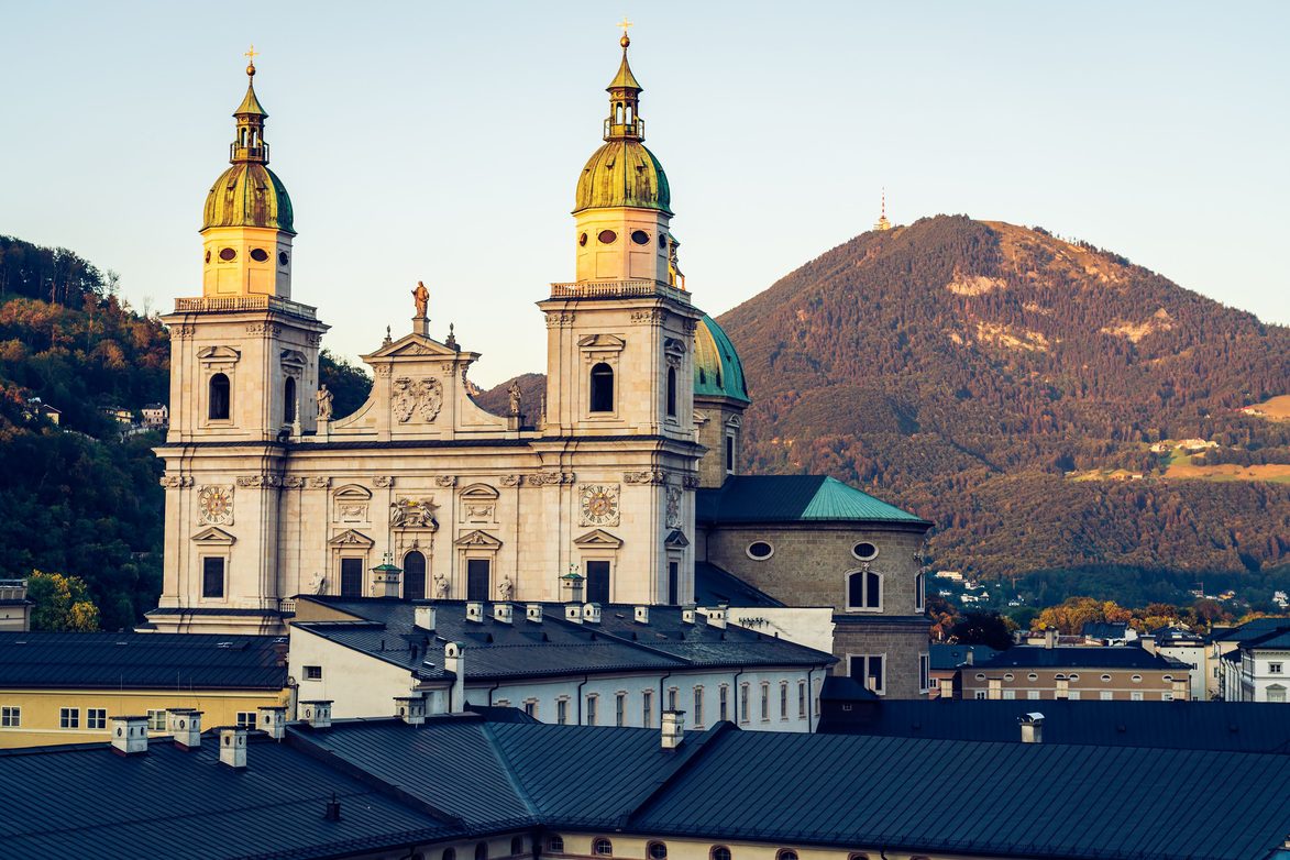 The pale marble facade and twin green-domed towers of Salzburg Cathedral, with the Gaisberg mountain behind