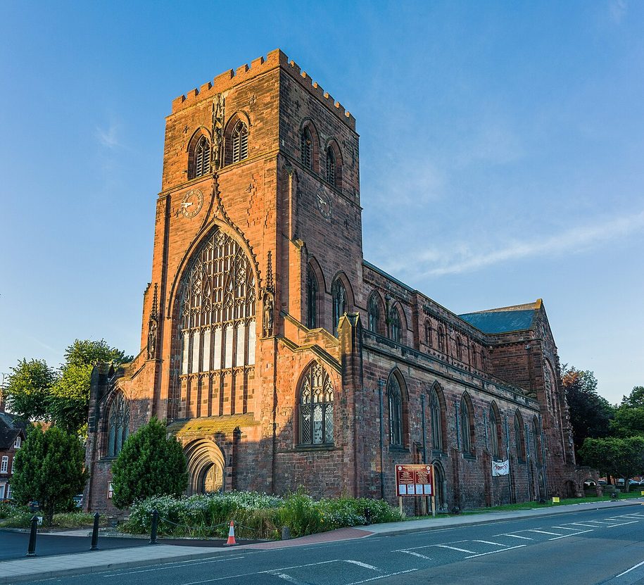 Shrewsbury Abbey in evening sunlight, its red sandstone west tower with a great Gothic window beside the long nave