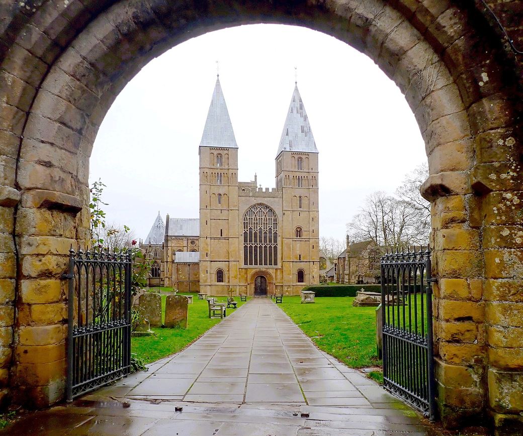 The west front of Southwell Minster framed by a stone gateway arch across the churchyard