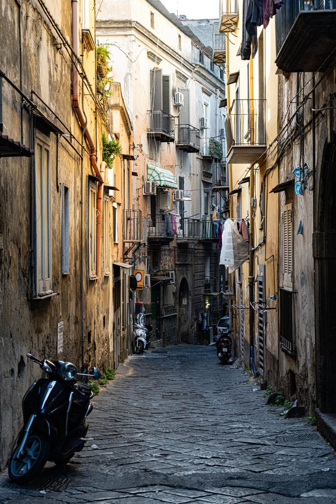 A narrow cobbled street in the historic centre of Naples, with tall old buildings and scooters parked along the walls