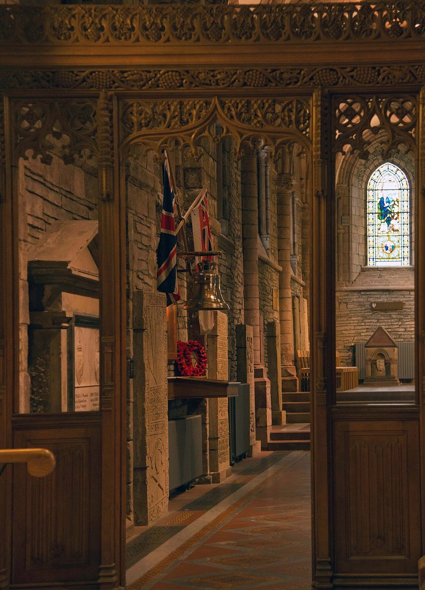 The HMS Royal Oak memorial in St Magnus Cathedral, with flags, a carved wooden screen and a stained-glass window