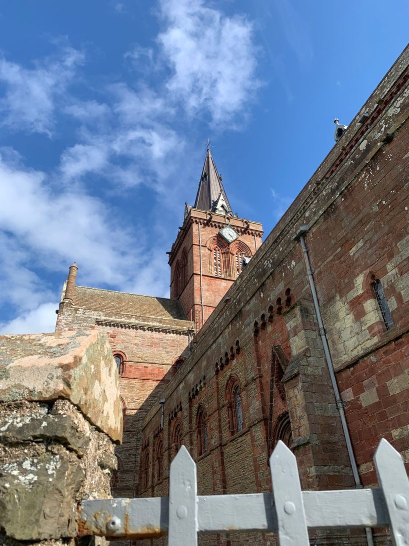 The long red and yellow sandstone side wall of St Magnus Cathedral with the tower and spire behind, under a blue sky