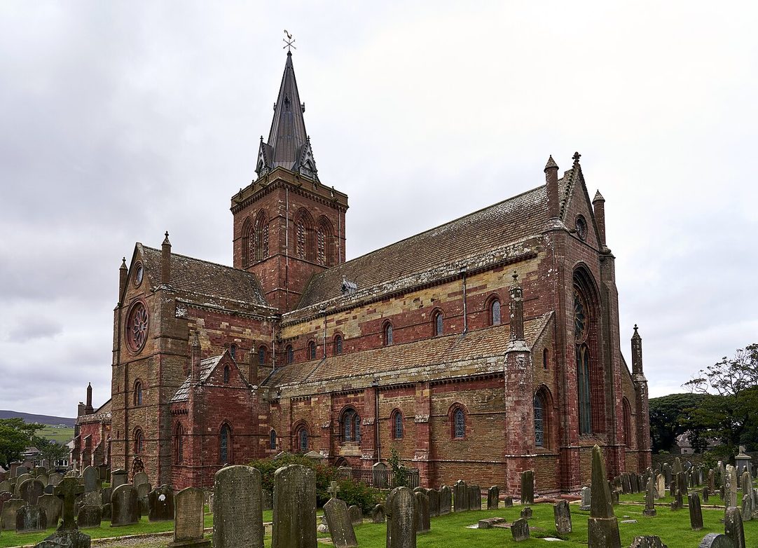 St Magnus Cathedral in Kirkwall, Orkney, a red sandstone Romanesque cathedral with a central tower and spire, seen across its graveyard