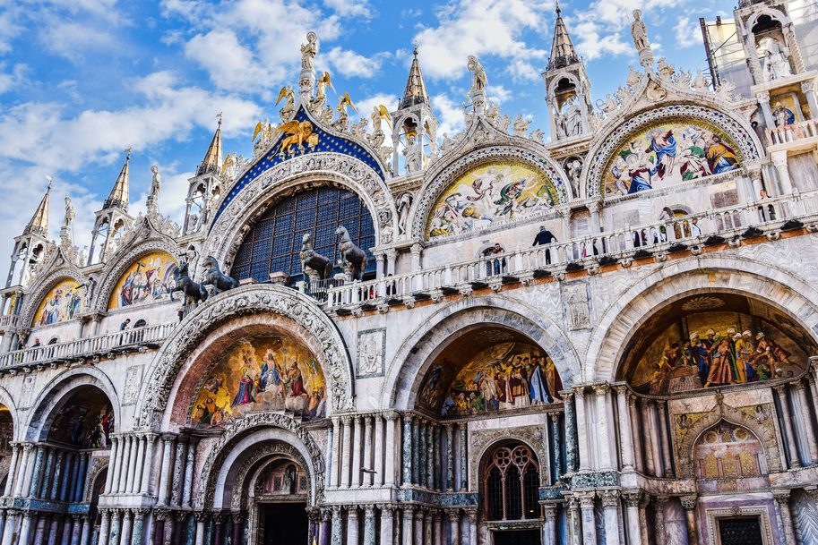 The west front of St Mark's Basilica in Venice, with gold mosaics in its arches and statues along the roofline