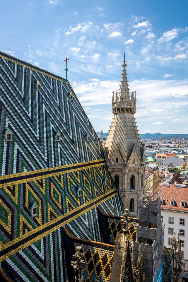 View down the patterned tiled roof of St Stephen's Cathedral from a tower, with a spire and Vienna beyond