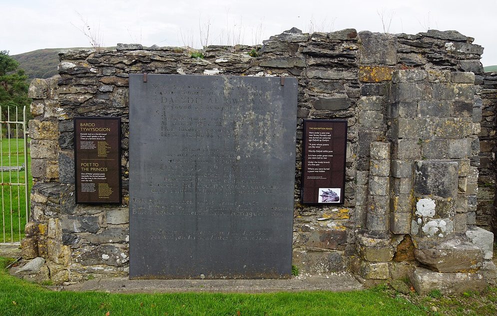 A dark stone memorial panel to the poet Dafydd ap Gwilym set against the ruined wall of the north transept at Strata Florida