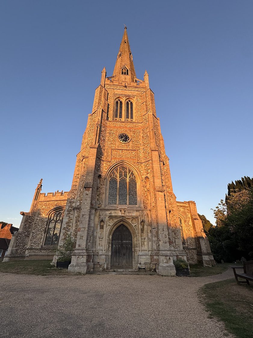 The west tower and spire of Thaxted church glowing orange in evening sunlight