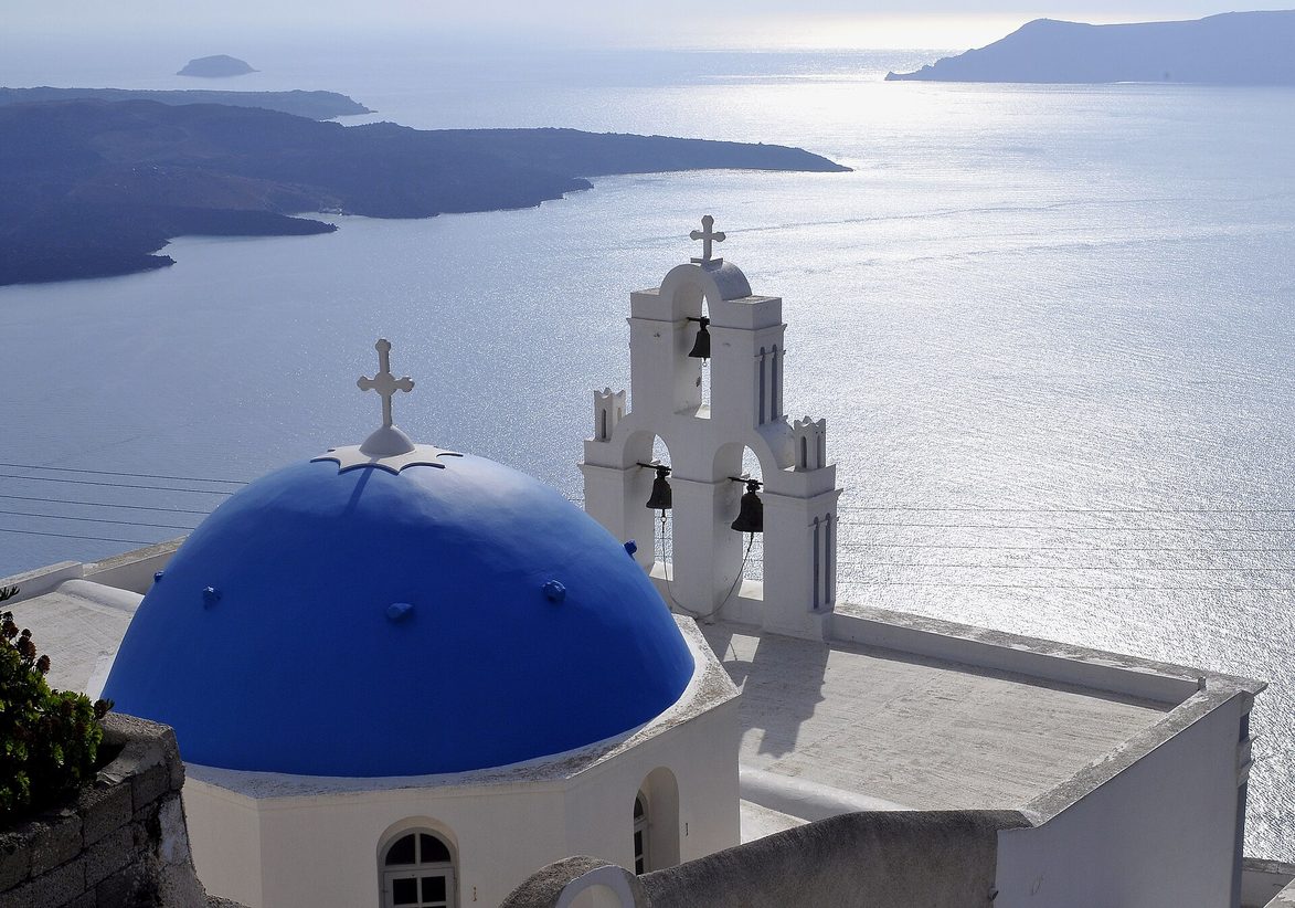 The blue dome and white bell gable of the Three Bells of Fira seen from the path above, with the caldera, the sea and the islands beyond